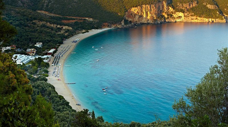 Lichnos beach from above: a long curve of sand with sunbeds beside turquoise water, below a golden cliff