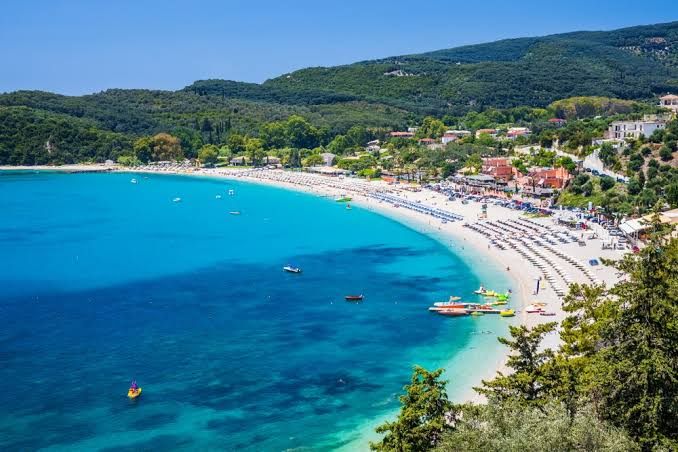 Valtos beach from above: a long curve of sunbeds and sand beside deep turquoise water, with green hills behind